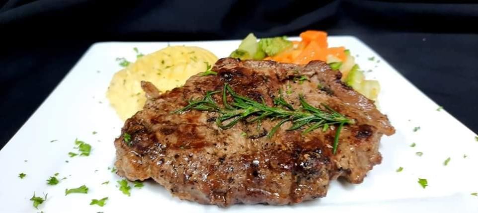 Imagen de un suculento churrasco cocinado al punto, dorado por fuera y jugoso por dentro, acompañado de una colorida variedad de vegetales frescos, que incluyen brócoli, zanahorias y pimientos. Además, el plato está servido con un delicioso puré de papas cremoso y suave. Los jugosos cortes de carne contrastan con los colores vibrantes de los vegetales y el puré, ofreciendo una combinación irresistible de sabores y texturas