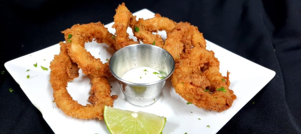 Imagen que muestra un plato de calamares de la casa empanizados, servidos con una deliciosa salsa tártara y una rodaja de limón fresco. Los calamares están crujientes por fuera y tiernos por dentro, con un dorado perfecto. La salsa tártara agrega un toque cremoso y sabroso, mientras que la rodaja de limón ofrece un toque cítrico y refrescante. Este plato es una combinación irresistible de texturas y sabores marinos