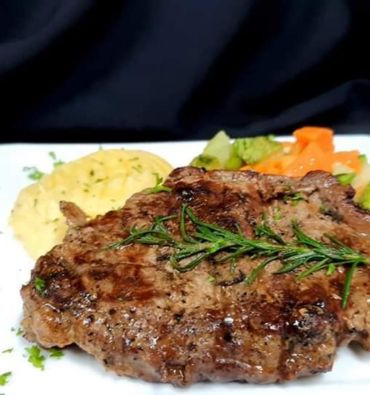 Imagen de un suculento churrasco cocinado al punto, dorado por fuera y jugoso por dentro, acompañado de una colorida variedad de vegetales frescos, que incluyen brócoli, zanahorias y pimientos. Además, el plato está servido con un delicioso puré de papas cremoso y suave. Los jugosos cortes de carne contrastan con los colores vibrantes de los vegetales y el puré, ofreciendo una combinación irresistible de sabores y texturas